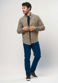 Beige Hemd mit Knöpfen aus strukturiertem Stoff, getragen über einem navyfarbigen T-Shirt, kombiniert mit blauen Jeans und braunen Schnürschuhen.