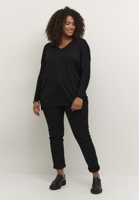 Schwarzer V-Ausschnitt-Pullover mit langen Ärmeln, lockere Passform, kombiniert mit schwarzen Jeans mit hochgekrempelten Säumen und glänzenden schwarzen Slippern vor einem neutralen Hintergrund.