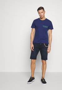 Marineblauwe t-shirt met groene tekst op de borst, gecombineerd met donkere denim shorts en zwarte sneakers met witte zolen. Eenvoudig ontwerp, casual pasvorm.