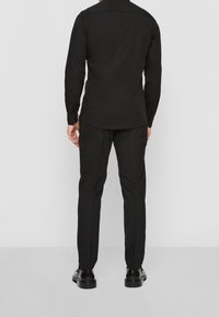 Chemise noire à manches longues avec un ourlet courbé, associée à un pantalon noir sur mesure et des chaussures noires à lacets, vue de dos.