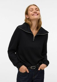 Svart hettegenser med glidelås, høy krage, lange ermer og ribbestrikkede mansjetter. Brukt med mørk denimjeans og svart belte. Glatt tekstur.