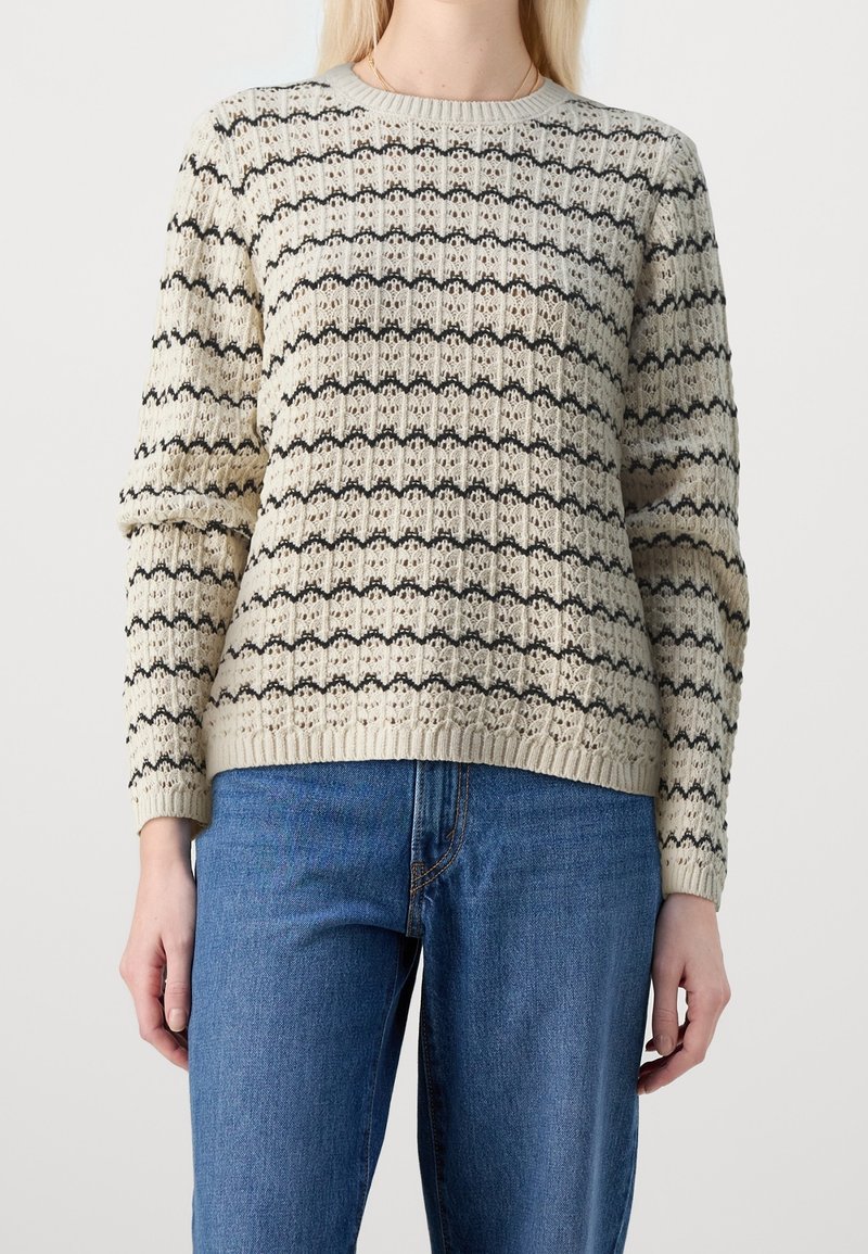 Personne portant un pull en maille crème avec des rayures horizontales noires et un jean bleu, debout devant un fond clair uni.