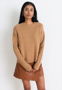 Femme aux longs cheveux noirs portant un pull en laine beige et une jupe en cuir marron, se tenant les mains jointes devant un fond blanc.