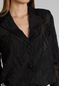 Zwarte textured blazer met ingewikkelde stofpatronen, een reverskraag en twee knopen aan de voorkant, met een semi-doorzichtige uitstraling.