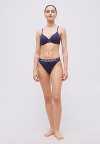 Γυναίκα όρθια, ξυπόλητη, φοράει μπλε σκούρο σουτιέν και αντίστοιχο εσώρουχο με το λογότυπο Polo Ralph Lauren, σε απλό φόντο.