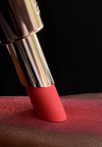 Krämig röd läppstift appliceras i en vinkel på strukturerad hud, vilket visar en mjuk, jämn applicering mot en mörk bakgrund.