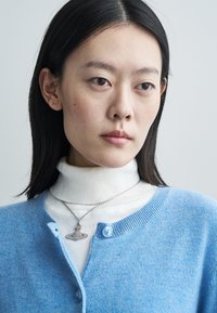 Light blue cardigan with round neck and button closure, layered over a white turtleneck. A silver necklace with a decorative pendant is worn.