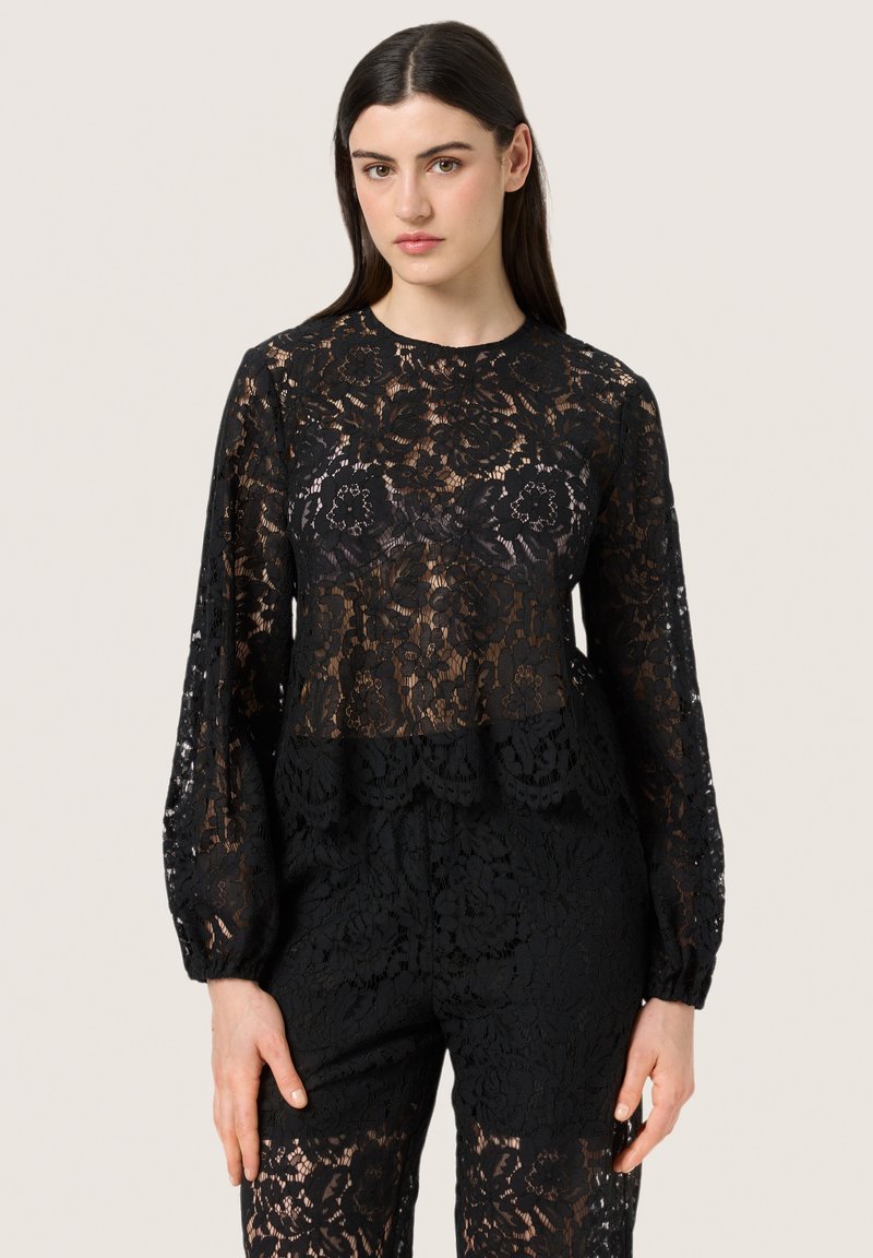 Top a maniche lunghe in pizzo nero con orlo scallop e motivo floreale, sovrapposto a un fondo in pizzo nero coordinato, con una texture semi-trasparente.