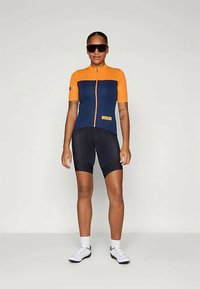 Īsām piedurknēm velosipēdu krekls tumši zilā krāsā ar oranžiem akcentiem, aprīkots ar priekšējo rāvējslēdzēju un logotipa uzlīmi, kopā ar melnām velosipēdu biksēm.