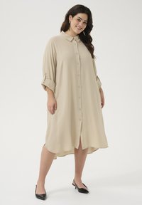 Beige langwerpige blousejurk met een knoopsluiting aan de voorkant, kraag, opgerolde mouwen en zijsplitten, gemaakt van lichtgewicht stof. Zwarte hakken.