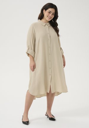 Beige long-sleeve shirt dress with a button front, collared neck, rolled sleeves, and side slits, made from lightweight fabric. Black heels.