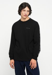 Černé tričko s dlouhým rukávem vyrobené z bavlny. Obsahuje malé logo na hrudi a text podél rukávů. Klasický kulatý výstřih.