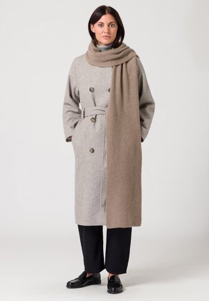 Manteau en laine gris avec un design double boutonnage, une taille ceintrée et de grands boutons, associé à une longue écharpe beige à côtes et des mocassins noirs.