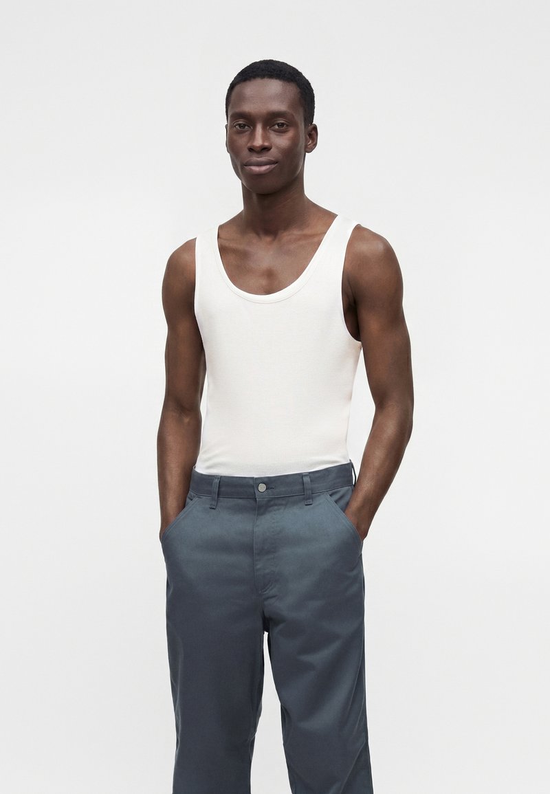 Jeune homme aux cheveux courts portant un débardeur blanc sans manches et un pantalon gris, debout les mains dans les poches, sur un fond uni.