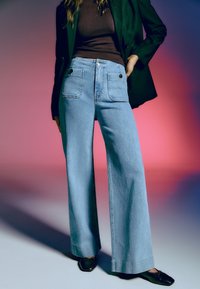 Femme portant un jean bleu clair à jambes larges avec poches plaquées à l'avant, un blazer noir, un haut marron et des chaussures noires à bout carré sur un fond rose et bleu.