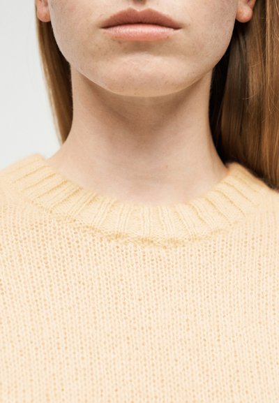 Gros plan sur le bas du visage et le cou d'une personne portant un pull tricoté jaune pâle avec un col rond côtelé.