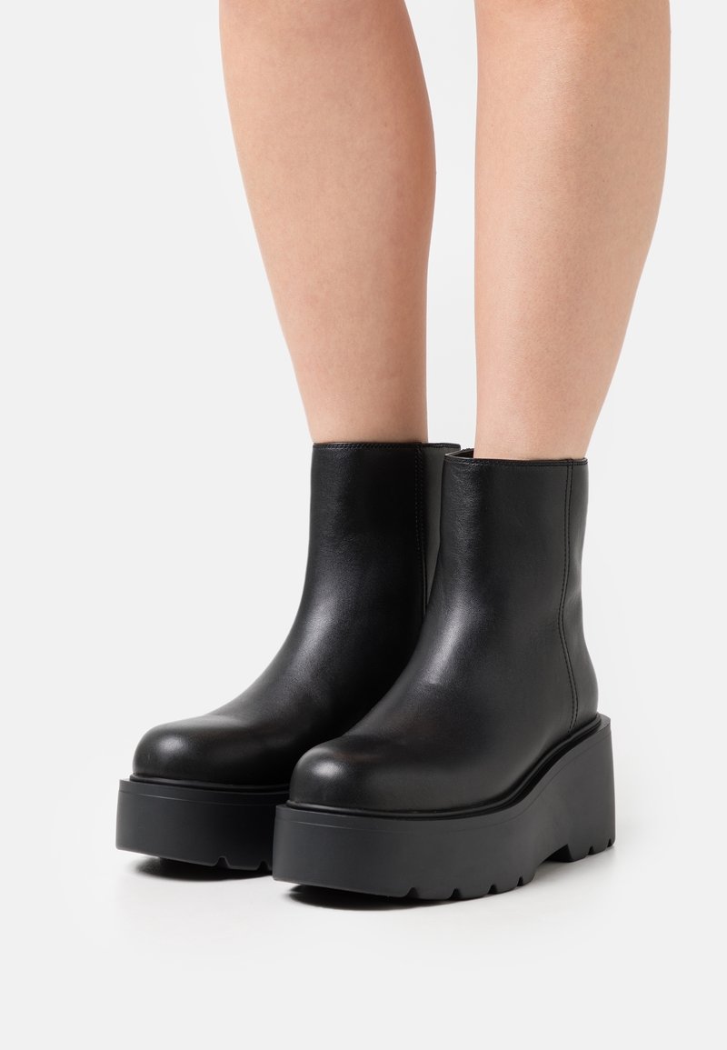 Bottines en cuir noir avec semelles épaisses à plateforme portées sur des jambes nues sur un fond clair uni.