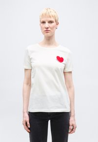 Άτομο με κοντά ξανθά μαλλιά φοράει λευκό T-shirt με κόκκινη καρδιά και το κείμενο «amour», συνδυασμένο με σκούρο τζιν, στέκεται μπροστά από απλό φόντο.