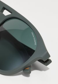Ochelari de soare din plastic gri cu lentile verzi. Prezintă un design geometric, text gravat "Armani Exchange" și accente subtile de portocalie pe brațe.