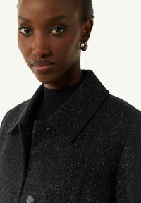 Black textured coat with a collared neckline and two large buttons. Worn over a ribbed black top, featuring a subtle sheen.