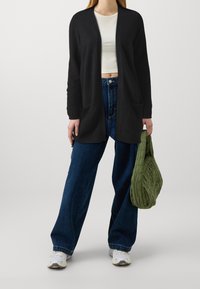 Svart cardigan med öppen framsida, ribbade ärmslut och två framfickor. Bärs över en vit crop top med hög midja och blå jeans samt en grön väska.