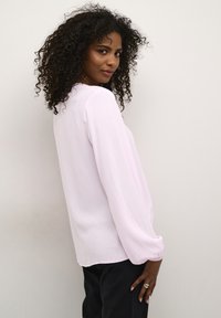 Haut rose pâle à manches longues avec un tissu texturé, col rond et poignets élastiqués, associé à un pantalon noir, vu de côté.