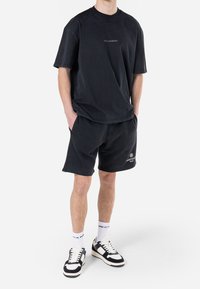 Černé oversized tričko s nápisem "PEGADOR", spárováno s černými kraťasy s detailním logem. Na nohou má černobílé tenisky a proužkované ponožky.