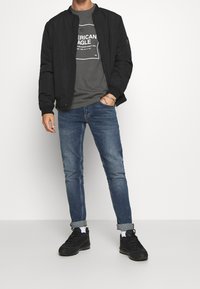 Svart bomberjacka, grå grafisk t-shirt, bleka blå jeans och svarta sneakers. Strukturerad tyg, avslappnad design, ledig passform.