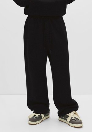 Person wearing loose black sweatpants and gray sneakers with thick beige laces, standing against a plain white background.