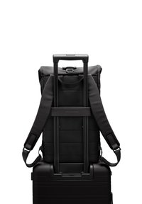 Sac à dos noir avec texture rembourrée, bretelles réglables et poignée rétractable, reposant sur une valise rigide noire.