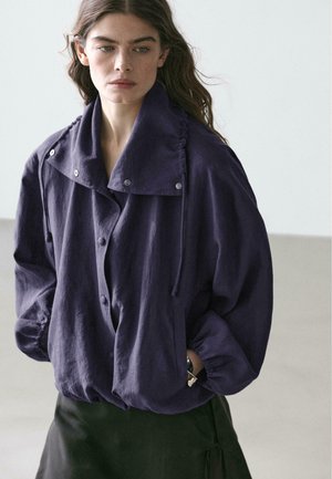 Donna con capelli lunghi castani indossa una giacca viola scuro larga con colletto grande ed elastico all'orlo, in piedi davanti a uno sfondo chiaro e semplice.