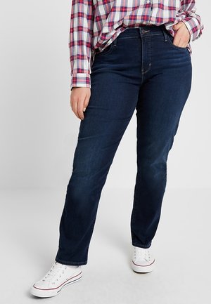 Person wearing dark blue jeans, white sneakers, and a red, white, and blue plaid shirt with one hand in pocket.