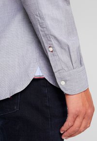 Vaaleansininen nappipaita teksturoidusta kankaasta, jossa on valkoiset napit ja punaiset tikkauskoristeet kalvosimissa sekä helman sivussa olevissa viilauksissa.