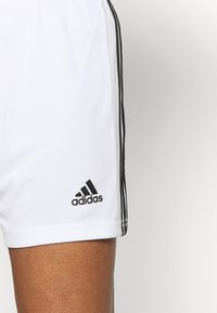 Vita Adidas-shorts med svarta tre-randsdetaljer längs sidan och Adidas-logotyp nära fållen på en persons lår.