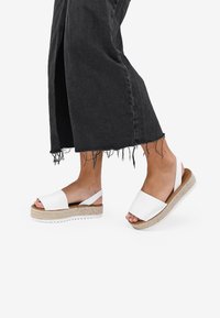 Sandalias de plataforma blancas con una correa ancha, plantilla de yute trenzada y suela de goma. Llevadas con pantalones negros de dobladillo deshilachado.