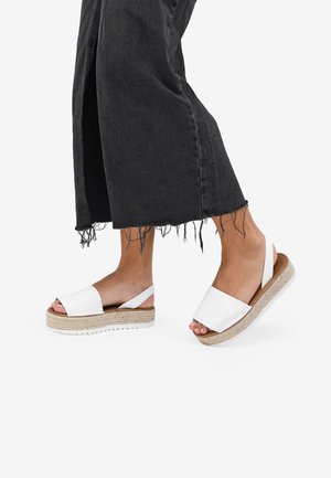 Sandalias de plataforma blancas con una correa ancha, plantilla de yute trenzada y suela de goma. Llevadas con pantalones negros de dobladillo deshilachado.
