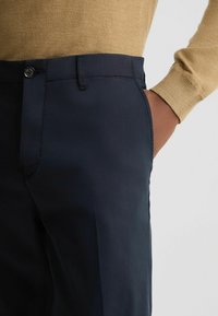 Marineblaufarbene Hose mit glatter Textur, diskreten vorderen Taschen und Knopfverschluss. Kombiniert mit einem hellbraunen, taillierten Strickpullover.