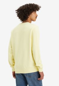 Man met krullend haar, gekleed in een lichtgele lange mouwen shirt en blauwe spijkerbroek, staand met zijn rug naar de camera tegen een witte achtergrond.