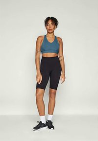 Mujer de pie con pantalones cortos de ciclista negros, sujetador deportivo color teal, calcetines blancos y zapatillas deportivas negras contra un fondo claro y liso.