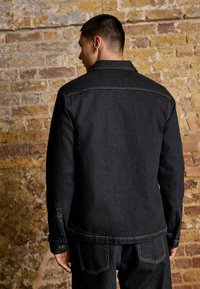 Black denim jacket with a collared neckline, visible stitching, and button cuffs. The design features a straight cut with rounded hem.