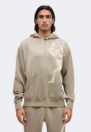 Mann mit langen dunklen Haaren trägt einen beigen Hoodie und passende Hose mit verblasster Basketballspieler-Grafik auf Brust und Oberschenkel.