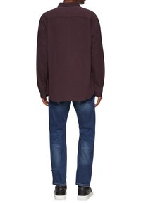 Chemise bordeaux à manches longues avec un col, détail au niveau du dos, et poignets boutonnés, associée à un jean en denim bleu et des baskets noires.