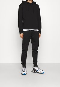 Schwarzer Hoodie mit Kordelzug-Kapuze, kombiniert mit passenden schwarzen Jogginghosen. Darunter ein weißes T-Shirt. Die Schuhe haben blaue Akzente.