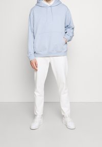 Sudadera de algodón azul claro con un bolsillo frontal, usada sobre una camisa blanca de botones, combinada con pantalones blancos ajustados y zapatillas blancas.