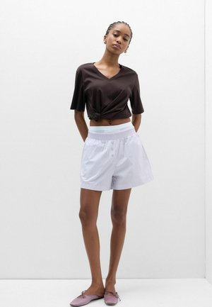 Frau trägt ein geknotetes schwarzes V-Ausschnitt-Top, weiße gestreifte Shorts und violette Ballerinas und steht vor einem schlichten weißen Hintergrund.