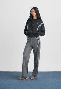 Melna jaka ar rāvējslēdzēju un baltām svītrām, pelēki sporta bikses ar sānu svītrām un melns t-krekls ar grafiku. Sporta apavi izceļ kopējo izskatu.