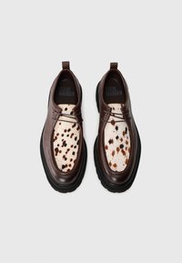 Pantofi din piele maro cu un model distinctiv al superiorului cu pete albe și maro, talpă din cauciuc neagră, design cu sireturi și un logo al mărcii în interior.
