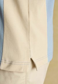 Close-up of a beige and light blue textured shirt hem with side slit over beige pants.