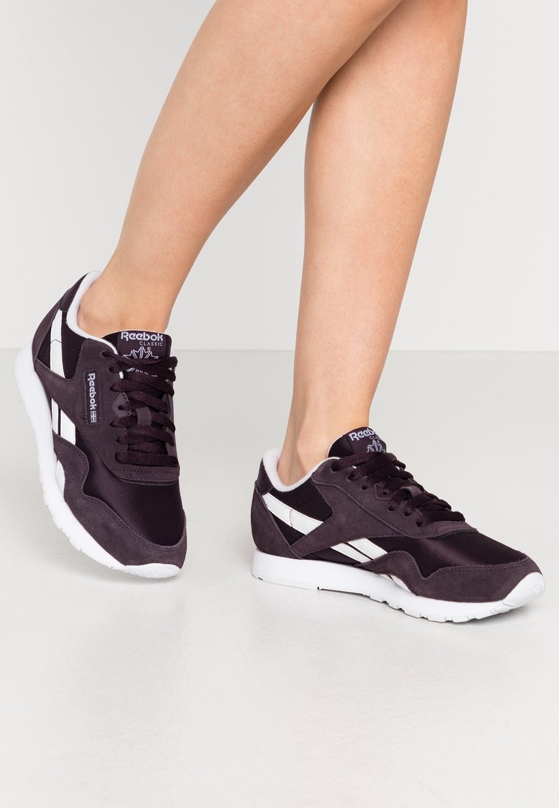 Jambes portant des baskets Reebok Classic noires et blanches avec des semelles blanches sur un sol et un fond gris clair.