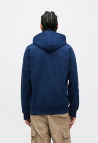 Tamsiai mėlynas džemperis su gaubtu, minkšta tekstūra, ribiniai rankogaliai ir apačia bei kengi stiliaus kišenė. Dėvimas su šviesiai chaki spalvos kargo kelnėmis.